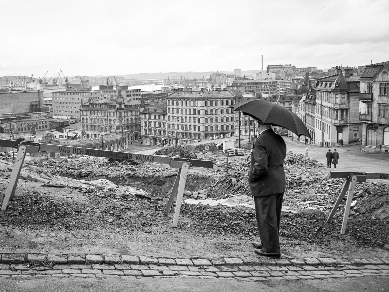 Mustavalkoinen valokuva, jossa tummaan pukuun pukeutunut mies tarkastelee kaupunkinäkymää sateenvarjon suojissa.