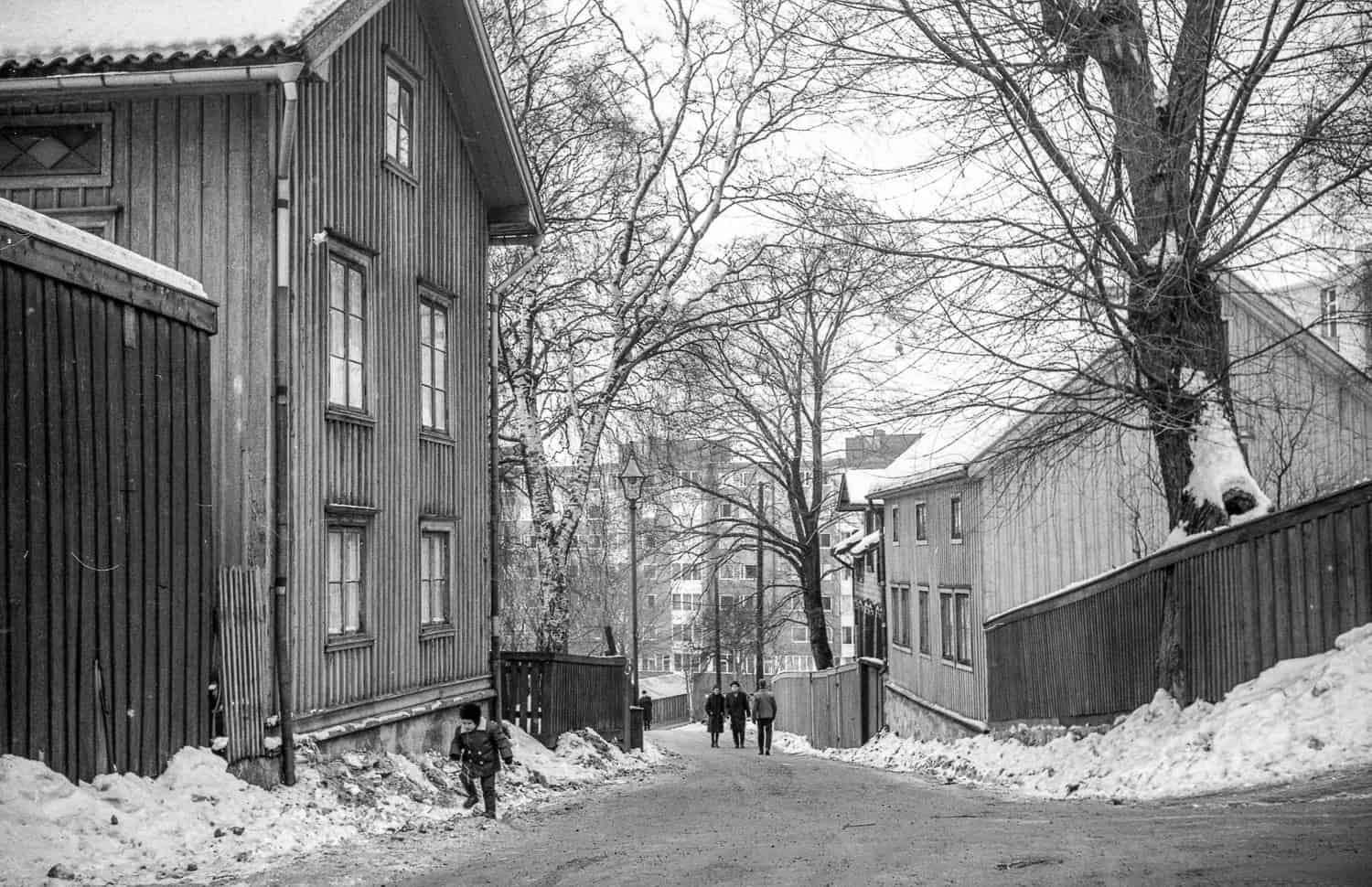 Mustavalkoinen valokuva kadusta, jonka varrella on puisia asuinrakennuksia. Kadun reunoilla on lumipenkkoja. Etualalla on lapsi lumipenkan lähellä, taaempana kadulla kävelee aikuisia ihmisiä.
