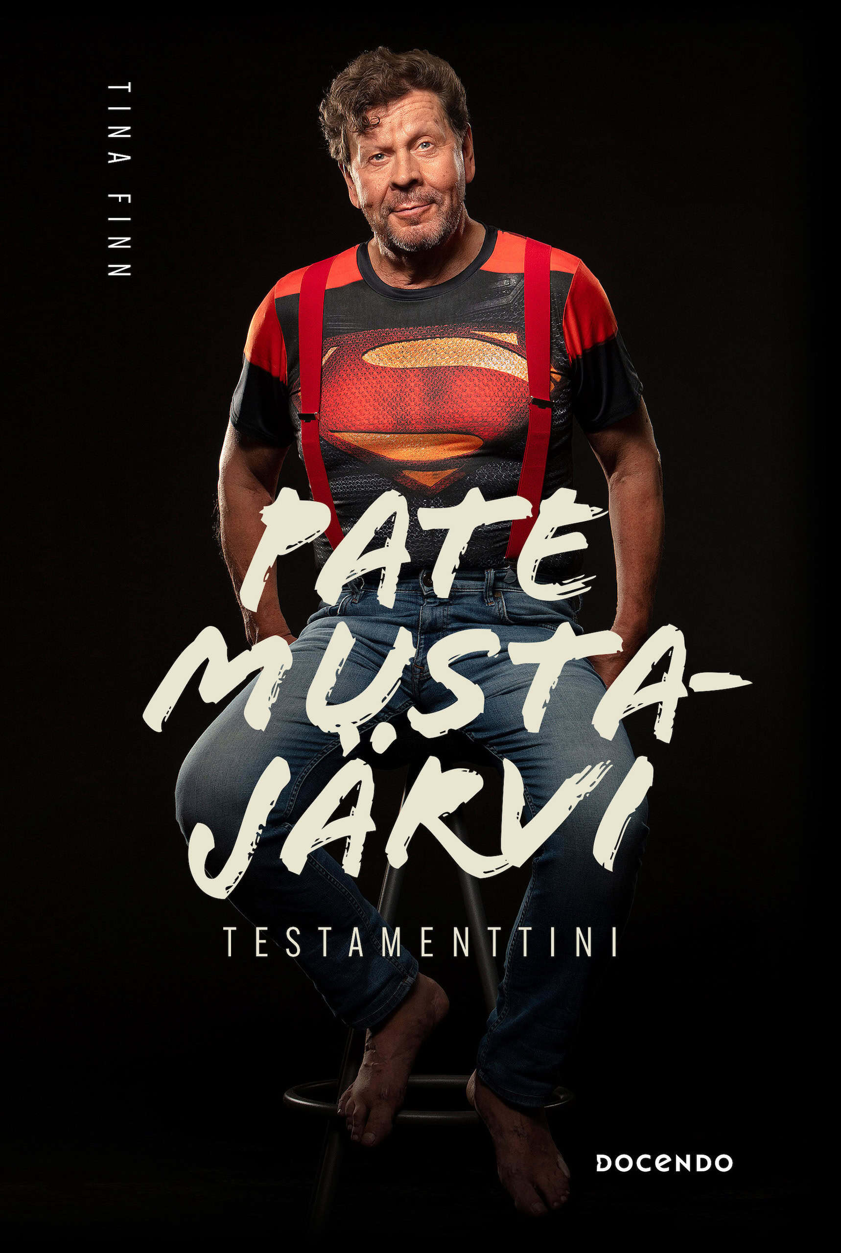 Testamenttini-kirjan kannessa on mustalla taustalla valokuva Pate Mustajärvestä istumassa tuolissa Teräsmies-paita päällään.