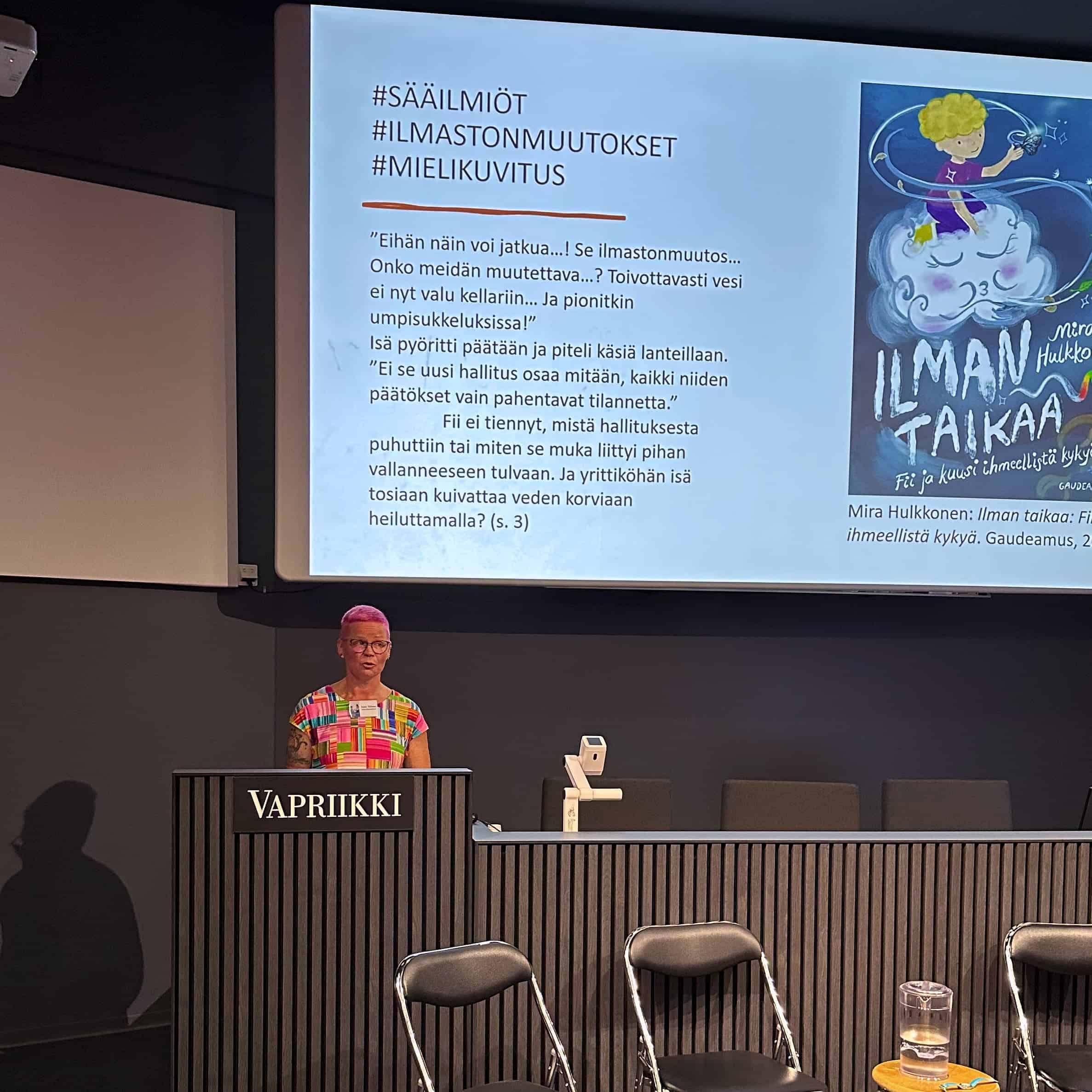 Lastenkirjainstituutin Anna Tiitinen Vapriikin auditoriossa, valkokankaalla dia Ilman taikaa -kirjasta.